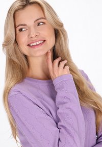 Lichtpaarse trui met een gestructureerd patroon, ronde hals en lange mouwen met kleine verhoogde stippen. Model raakt haar nek aan.