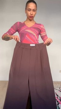 Person wearing a pink patterned top holds wide brown pants in front of a plain wall and light floor indoors.