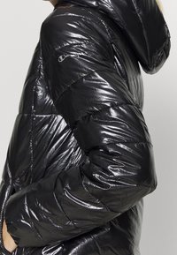 Svart pufferjacka med glänsande yta, har huva, quiltat mönster och logotyp på ärmen. Texturerad tyg med smidig silhuett.