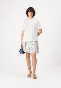 Weißes T-Shirt mit Fransen, kombiniert mit einem Rock mit Blumenmuster. Dazu braune Riemchensandalen und eine kleine blaue Handtasche.