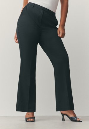 Donna con pantaloni neri a vita alta e gamba dritta e sandali neri con tacco e punta aperta, in piedi davanti a uno sfondo semplice.