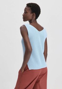 Blauwe gebreide mouwloze top met een V-hals, textuurstof en open rugontwerp, gecombineerd met losse roestkleurige broek.