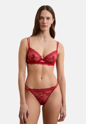 Frau trägt roten Spitzen-BH und passende Spitzen-Pantys, steht vor einem schlichten weißen Hintergrund, blickt nach vorne mit neutralem Ausdruck.
