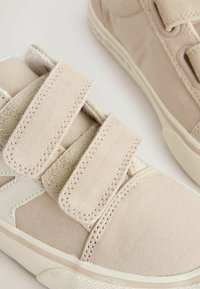 Beige Stoff-Sneaker mit zwei Klettverschlüssen, strukturierten Wildleder-Akzenten und einer Gummisohle mit einem subtilen Fischgrätenmuster.