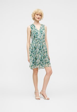 Ung kvinde med kort krøllet blondt hår, iført en ærmeløs, knælang kjole med grønne og blå mønstre samt hvide højhælede sandaler.