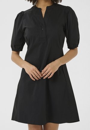 Femme portant une courte robe noire avec des manches courtes bouffantes et un bouton unique au col en V, les mains doucement jointes à la taille.