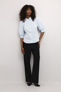 Camicia azzurra con maniche a palloncino e bottoni sul davanti, rifinita in pizzo, abbinata a pantaloni neri su misura e scarpe nere con punta.