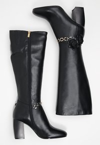 Czarne skórzane kozaki za kolano na słupku, z suwakiem z przodu i ozdobnym łańcuszkiem z kwiatowymi akcentami po boku. Gładka faktura.