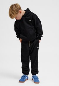 Schwarzer Hoodie mit einer Vorderentasche und Logo auf der Brust, kombiniert mit schwarzen Jogginghosen und blauen Sneakers mit Klettverschlüssen.