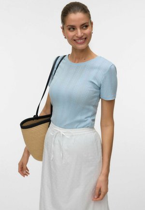 Smiling woman wearing light blue short-sleeve top, white skirt with drawstring, carrying woven straw tote bag with black straps over shoulder.