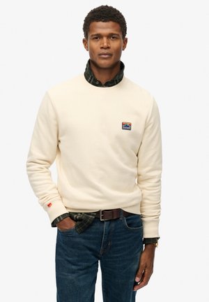 Crème sweatshirt met een ronde hals en een klein kleurrijk berglogo, gelaagd over een donkergroene geruite shirt, gecombineerd met blauwe jeans.