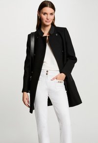 Cappotto nero di lana con colletto alto, bottoni doppi e dettagli testurizzati. Abbinato a pantaloni bianchi con hardware dorato.