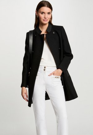 Black wool coat with a high collar, double-breasted buttons, and textured accents. Paired with white pants featuring gold hardware.