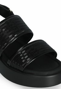 Sandales noires avec des brides ornées de sequins, une semelle plate et une finition mate et lisse. Conçues pour le confort avec un look épuré et minimaliste.