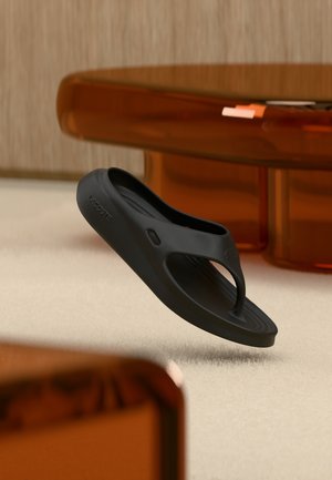 Infradito nero in gomma, con design a perno, suola con texture e dettaglio logo in rilievo. Posto su una superficie morbida e chiara.