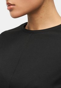 Κοντινό πλάνο του κάτω μέρους του προσώπου και του λαιμού ενός ατόμου που φοράει μαύρο μπλουζάκι με στρογγυλή λαιμόκοψη, μπροστά σε ένα απλό φόντο.