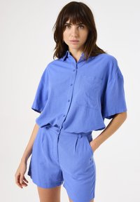 Camisa de manga corta azul claro con cuello, bolsillo frontal y cierre de botones, combinada con pantalones cortos de cintura alta a juego. Textura suave.