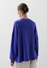 Oversized pullover in een levendige blauwe stof, met verlaagde schouders, ronde hals en een gestructureerde afwerking, gezien vanaf de achterkant.