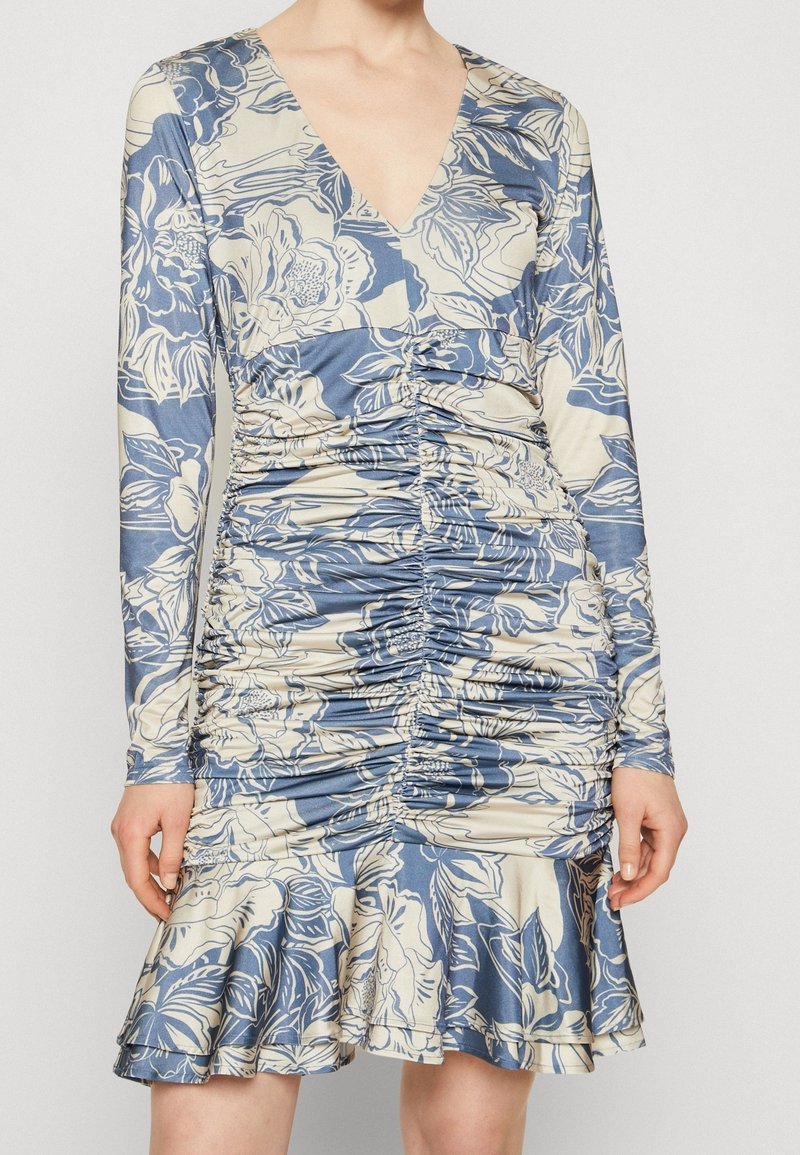 Robe florale à manches longues et ruchée en bleu et beige avec un décolleté en V et un ourlet volanté portée par une personne avec les mains détendues sur les côtés.