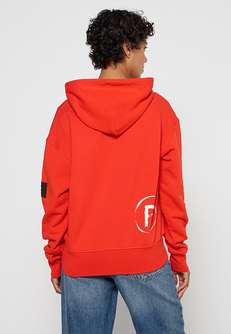 Czerwona bluza z kapturem z kieszenią typu kangur na przodzie, biały okrągły logo z tyłu i czarny akcent na lewym rękawie. Miękki, polarowy materiał.