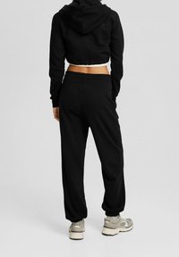 Svart kort hoodie med huva, kombinerad med svarta joggers. Båda har en avslappnad passform och elastiska muddar. Bärs med ljusa sneakers.