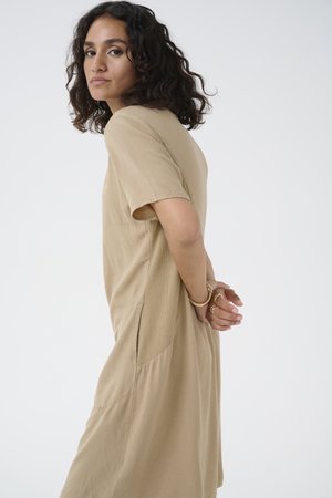 Short-sleeve beige dress with a relaxed fit, textured fabric, and side pockets. Model has curly hair and hands clasped behind back.