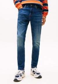 Mees, kes kannab siniseid kitsaid teksapükse, valgeid ja mereväe spordijalatsid ning pikade varrukatega oranži ja mereväe triibulist särk, seisab valge tausta ees.