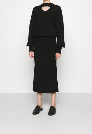 Person iført en sort ribstrikket sweater med udskåret halsudskæring og matchende nederdel i midi-længde, kombineret med sorte slip-on loafers.