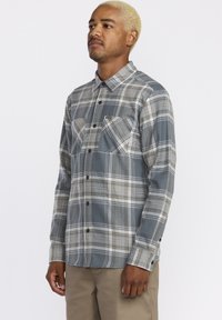 Chemise en flanelle à carreaux gris, blanc et beige. Présente un devant à boutons, deux poches poitrine et des manches longues avec des poignets à boutons.
