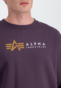 Ljubičasti dukserica s okruglim izrezom i zlatnim grafičkim logotipom s natpisom "ALPHA INDUSTRIES". Mekani materijal s glatkom teksturom.