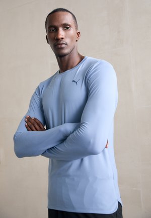 Homme les bras croisés portant un t-shirt à manches longues Puma bleu clair, debout contre un mur beige uni.