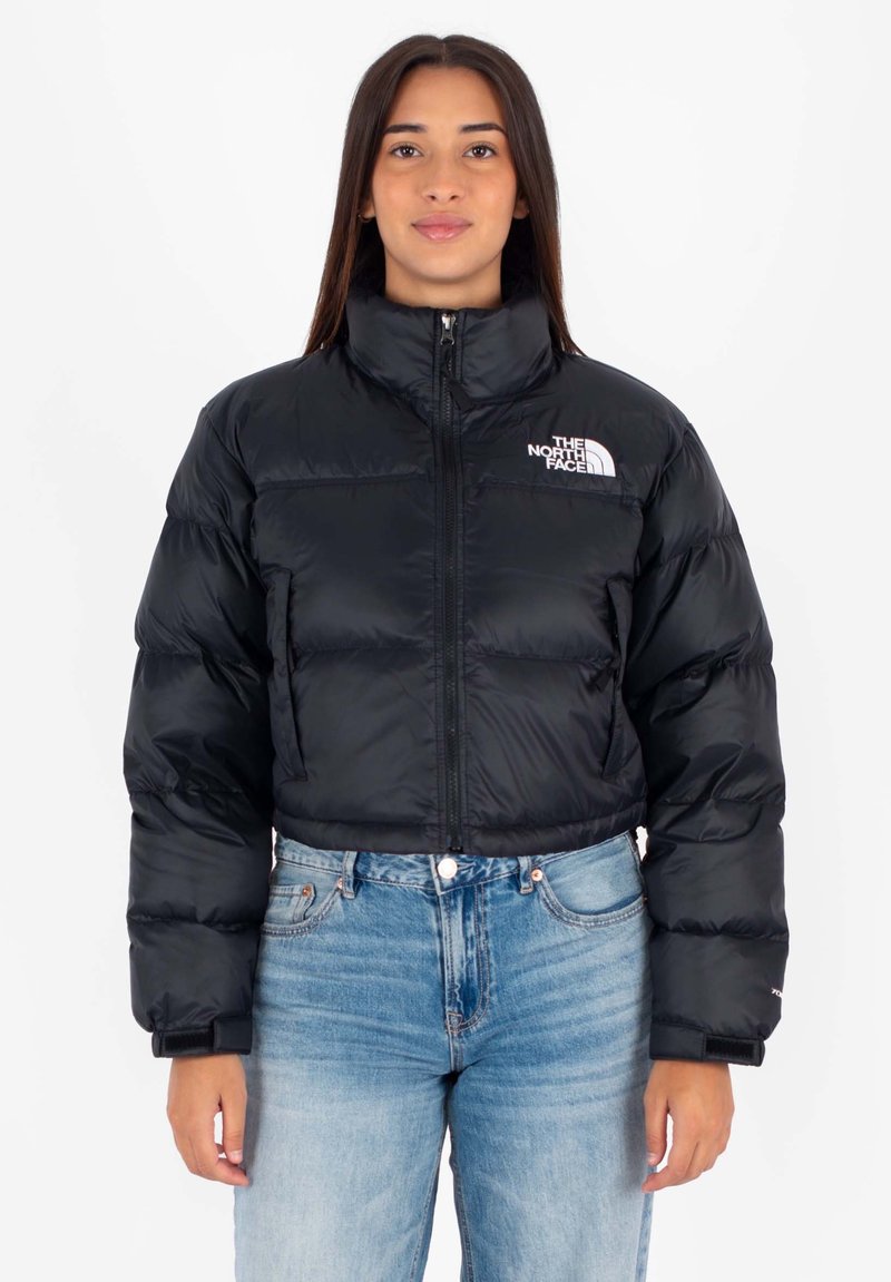 Giovane donna che indossa un giubbotto corto imbottito nero con il logo The North Face e jeans alta vita azzurri, rivolta frontalmente.