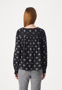 Femme aux cheveux rouges lisses portant un chemisier noir à manches longues avec des motifs floraux et un jean gris, vue de dos sur un fond blanc.