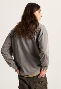 Šedá dlouhá košile s texturovaným designem na zadní straně, doplněná o tmavé cargo kalhoty s potiskem. Detaily zahrnují stříbrné šperky.