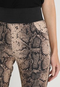 Leggings de estampado animal que presentan una cinturilla negra ajustada y un fondo texturizado en tono marrón claro con patrones oscuros similares a serpientes y un ligero brillo.