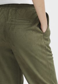 Olivenfargede, myke joggebukser i suedelignende materiale med elastisk midje, sidelommer og en enkelt baklomme. Har en jevn tekstur.