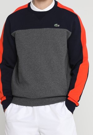 Mann trägt ein graues und marineblaues Sweatshirt mit leuchtend orangefarbenen Streifen an den Ärmeln und einem kleinen grünen Krokodil-Logo auf der Brust.