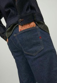 Un jean en denim bleu foncé présente des coutures orange contrastantes, une poche arrière distinctive avec un accent rouge et un design près du corps.