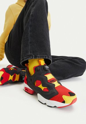 Osoba nosząca czerwono-żółto-czarne buty Reebok, żółte skarpetki, czarne dżinsy i żółtą bluzkę z długim rękawem, siedząca ze skrzyżowanymi nogami.