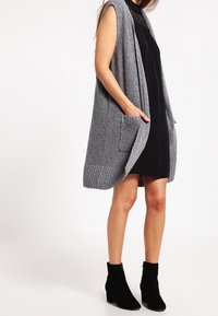 Szary dzianinowy bezrękawnik z kieszeniami, otwartym przodem i prążkowanym dołem, noszony na czarnej sukience i zestawiony z czarnymi botkami za kostkę.