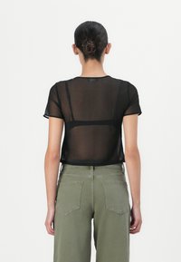 Top nero trasparente a maniche corte con scollatura rotonda, abbinato a pantaloni olive verdi a vita alta, che mettono in risalto un tessuto trasparente.
