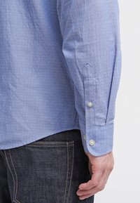 Vaaleansininen napitettava paita, jossa on pieni, toistuva geometrinen kuvio. Siinä on suora helma ja napilliset kalvosimet. Päällä tummat farkut.