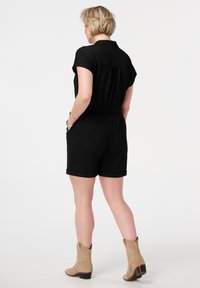 Combinaison à manches courtes noire avec col, taille élastique et poches latérales. Fabriquée en tissu texturé, associée à des bottines couleur brun clair.