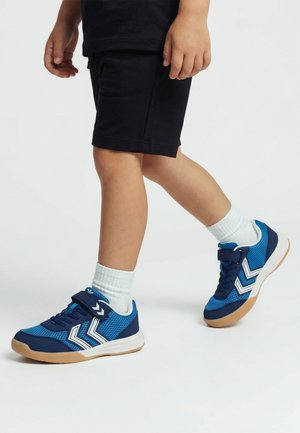 Dziecko w czarnych szortach i białych skarpetkach chodzące w niebieskich i granatowych sneakersach z białymi paskami w jodełkę na jasnym tle.