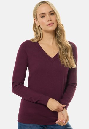 Maglione a V in un profondo colore prugna, realizzato in morbido tessuto knit con maniche lunghe e polsini a coste, indossato sopra dei jeans. Design ordinato e aderente.