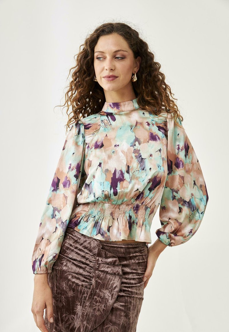 Blusa de estampado floral con cuello alto, mangas abullonadas y cintura fruncida. Los colores incluyen verde menta, durazno y púrpura sobre una tela suave.