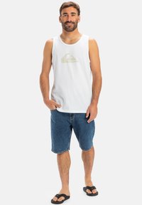 Hvit bomullstopp med lys beige logo, kombinert med blå denimshorts og svarte flip-flops. Uformell sommerantrekk.