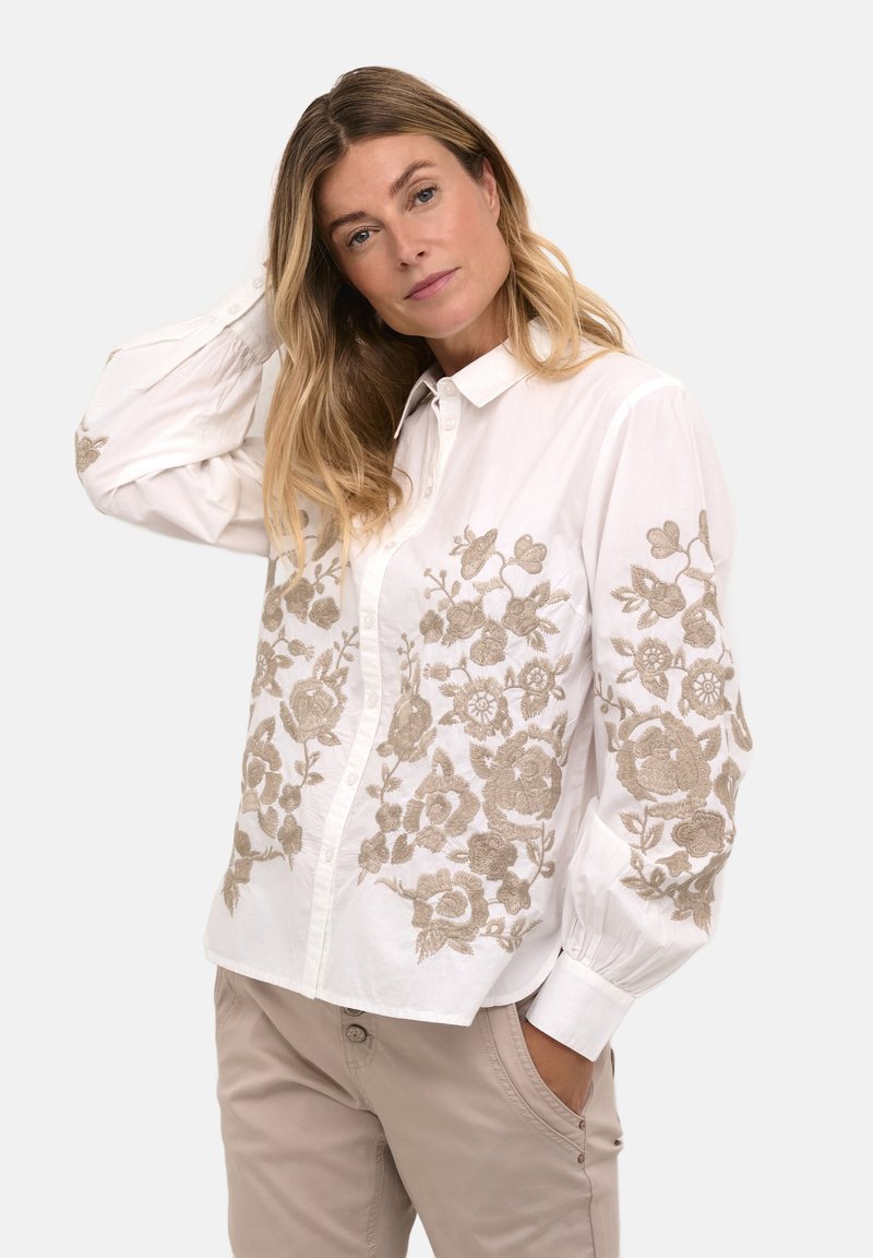 Cămașă albă cu nasturi, având broderie florală complicată în nuanțe de bej pe față și mâneci; realizată dintr-un material ușor, cu o textură netedă.