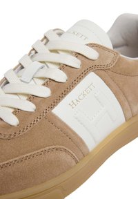Bruna mockainsneakers med vita läderdetaljer, med en framträdande präglad 'H'-logotyp, vita snören och en lätt gummisula.