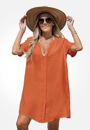 Žena si priaže široký slamenný klobúk, má na sebe prefarbené oranžové šaty s krátkymi rukávmi, slnečné okuliare, viacvrstvové náhrdelníky a náramky, usmieva sa.